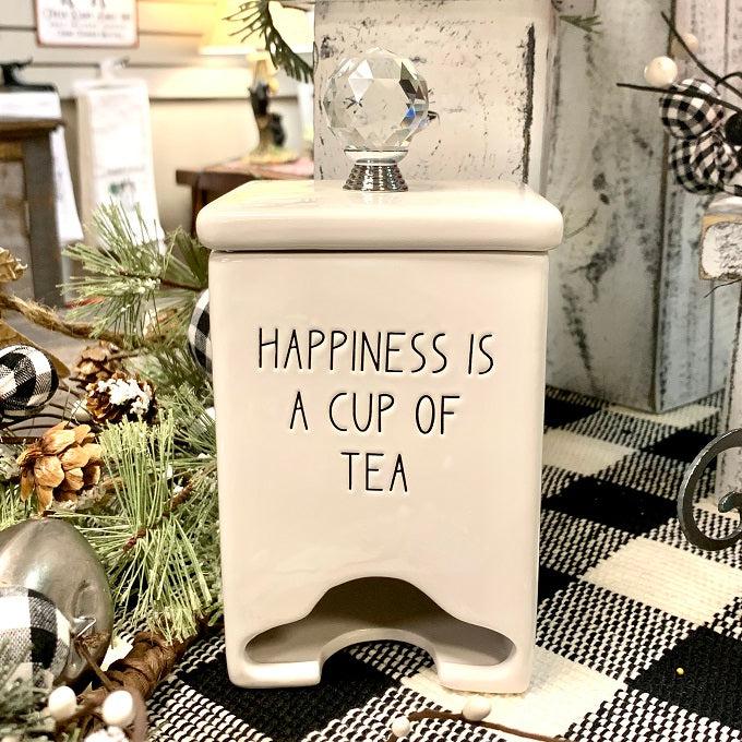 A white ceramic square tea bag dispenser with a removable lid that has a glass knob on the top. The saying on the front side of the dispenser is Happiness is a Cup of Tea.