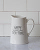A white ceramic pitcher with the words Happy Everything on the side in black. There is small black florals between the words. It is empty demonstrating that is could be used a juice or water pitcher.