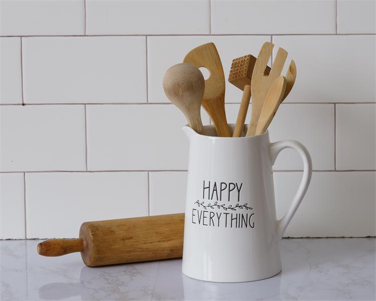 A white ceramic pitcher with the words Happy Everything on the side in black. There is small black florals between the words. It is filled with wooden utensils.