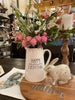 A white ceramic pitcher with the words Happy Everything on the side in black. There is small black florals between the words. It is filled with seasonal coloured florals.