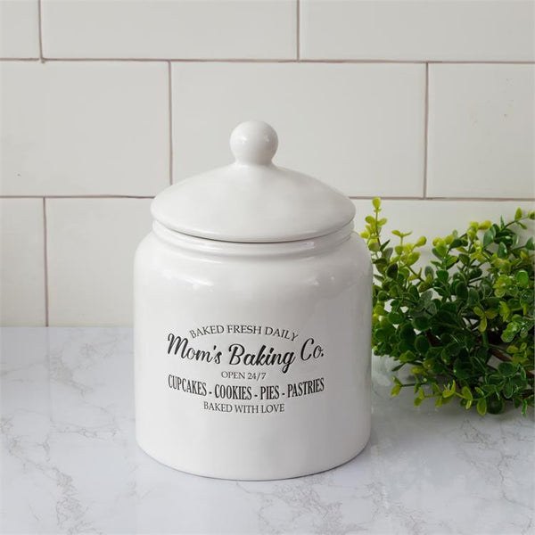 White ceramic cookie jar with lid is pictured. This farmhouse style jar has Mom's Baking Co. open 24/7, baked fresh daily imprinted on the side. It has a white removable lid.