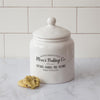 White ceramic cookie jar with lid is pictured. This farmhouse style jar has Mom's Baking Co. open 24/7, baked fresh daily imprinted on the side. It has a white removable lid.