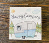 A small square wooden sign with a water-colour painting of a old-style camper. The camper is painted blue on the bottom and light gray on the top. There are evergreen trees in the background and a sun in the sky. The words Happy Campers is in script in the sky, above the camper.