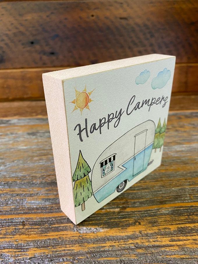 A side view showing the width of a small square wooden sign with a water-colour painting of a old-style camper. The camper is painted blue on the bottom and light gray on the top. There are evergreen trees in the background and a sun in the sky. The words Happy Campers is in script in the sky, above the camper.