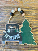 A farmhouse vintage black truck is packed with presents and beside a solid green evergreen tree on this wooden ornament with a beaded hanger. The NOEL banner is hanging across the back of the truck.