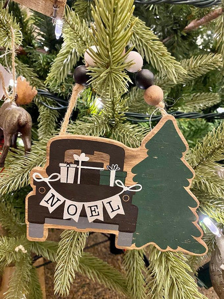 A farmhouse vintage black truck is packed with presents and beside a solid green evergreen tree on this wooden ornament with a beaded hanger. The NOEL banner is hanging across the back of the truck.