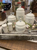 Grouping of white canisters in a decorative Tray. There are four white ceramic canisters , one for coffee, one for flour, one for sugar and one for tea. All have glass knobs. There is also a white set of salt and pepper shakers.
