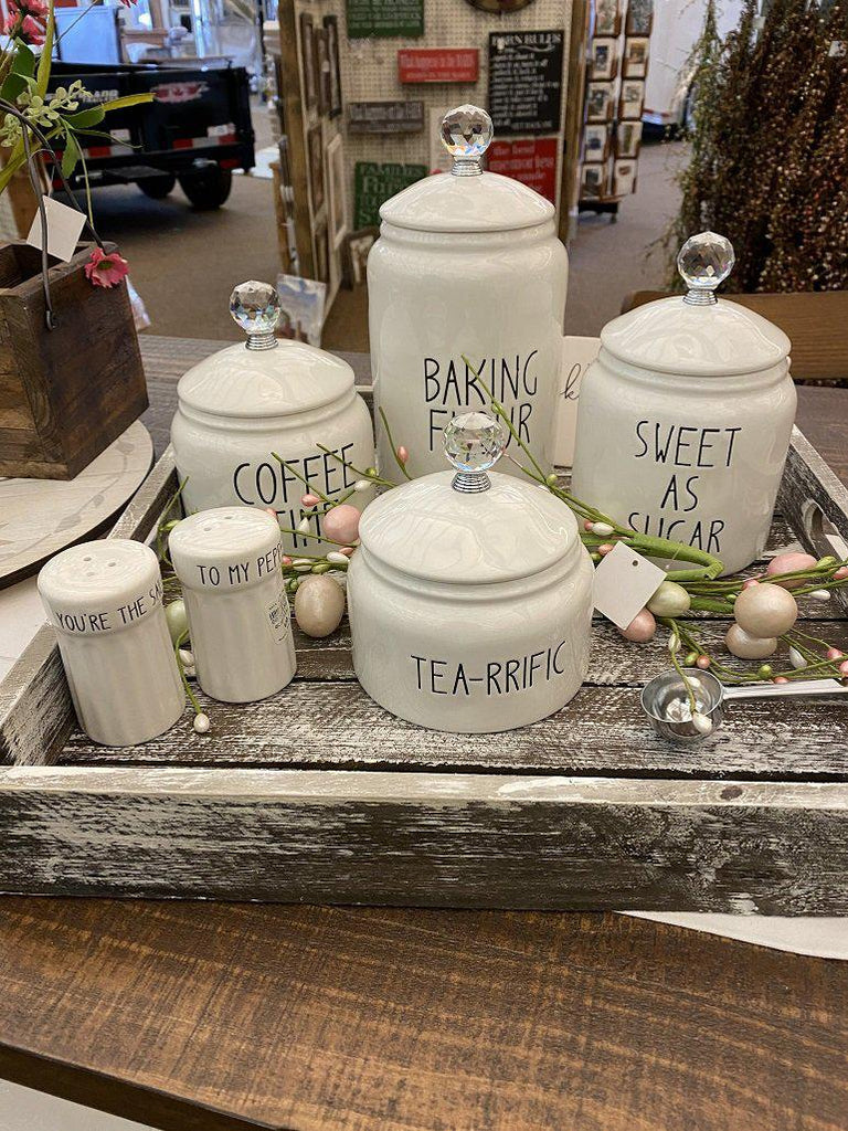 Grouping of white farmhouse-style  ceramic kitchen canisters in a decorative Tray. There is four white ceramic canisters , one for coffee, one for flour, one for sugar and one for tea. All have glass knobs. There is also a white set of salt and pepper shakers.. 