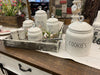 Grouping of white farmhouse-style ceramic kitchen canisters in a decorative Tray. There is five white ceramic canisters , one for coffee, one for flour, one for sugar, one for cookies and one for tea. All have glass knobs on the lids. There is also a white set of salt and pepper shakers.. 