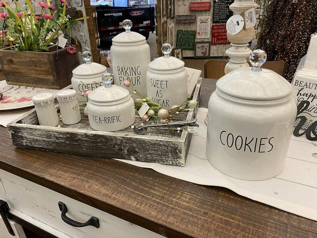 Grouping of white farmhouse-style ceramic kitchen canisters in a decorative Tray. There are five white ceramic canisters , one for coffee, one for flour, one for sugar, one for cookies, and one for tea. All have glass knobs on the lids. There is also a white set of salt and pepper shakers.