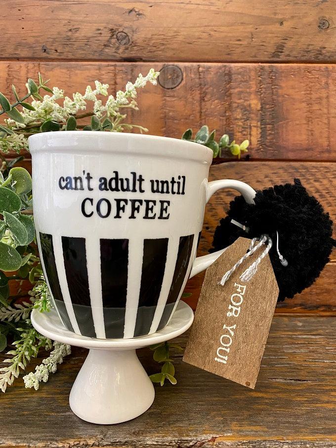 A white ceramic mug ready for gifting with a wooden gift tag and a black pom pom. The bottom half of the mug has black stripes and printed on both sides are the words can't adult until coffee.