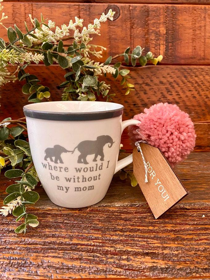 A white ceramic mug with a gray rim, pink pom pom and wooden gift tag. The mug is printed on both sides and has an image on a mother and infant elephant. The mug says where would I be without my mom.