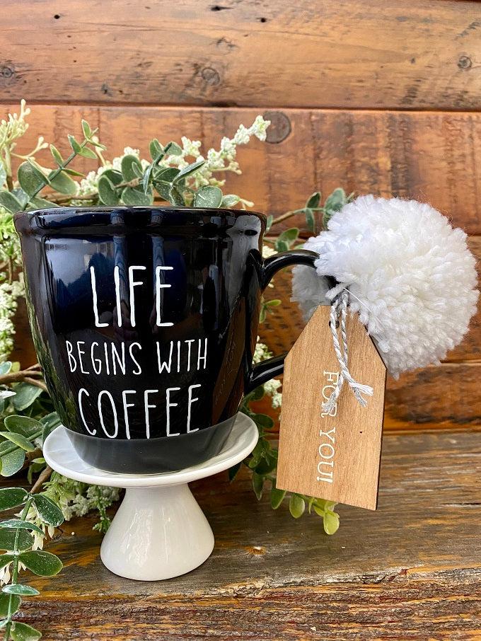 A black ceramic coffee mug printed on both sides with the words Life begins with Coffee. It comes ready for gifting with a pom pom and wooden gift tag.
