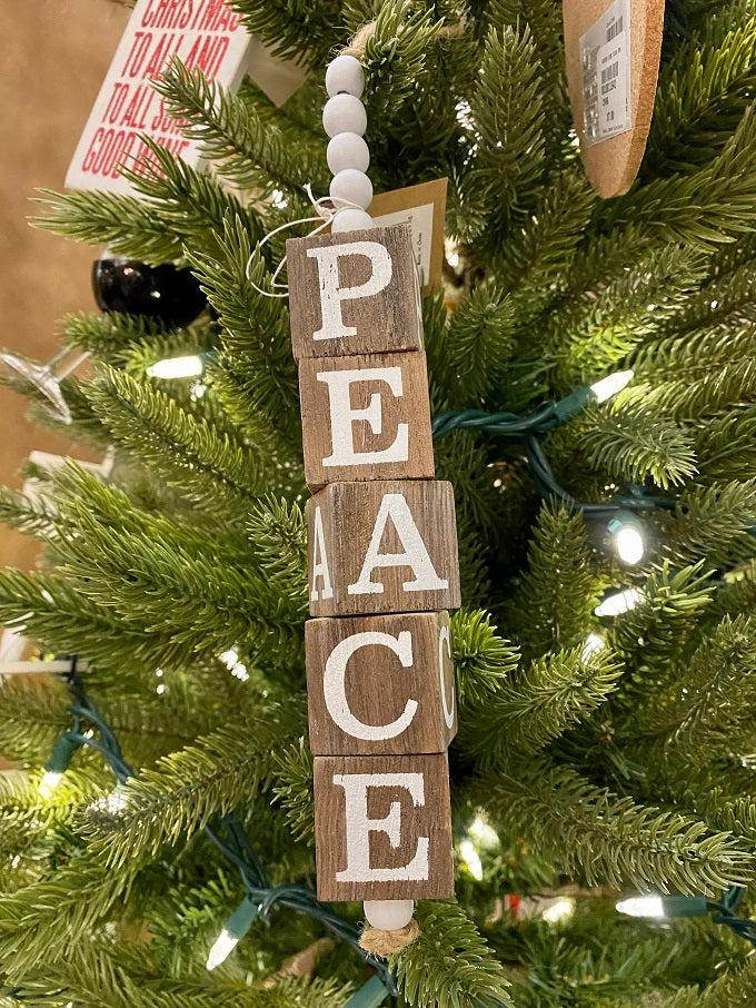 The Beaded Cube Ornament with the word Peace. The natural coloured cubes are printed with a single letter on all four sides and are moveable. Five white beads are at the top and one at the bottom of the ornament. 