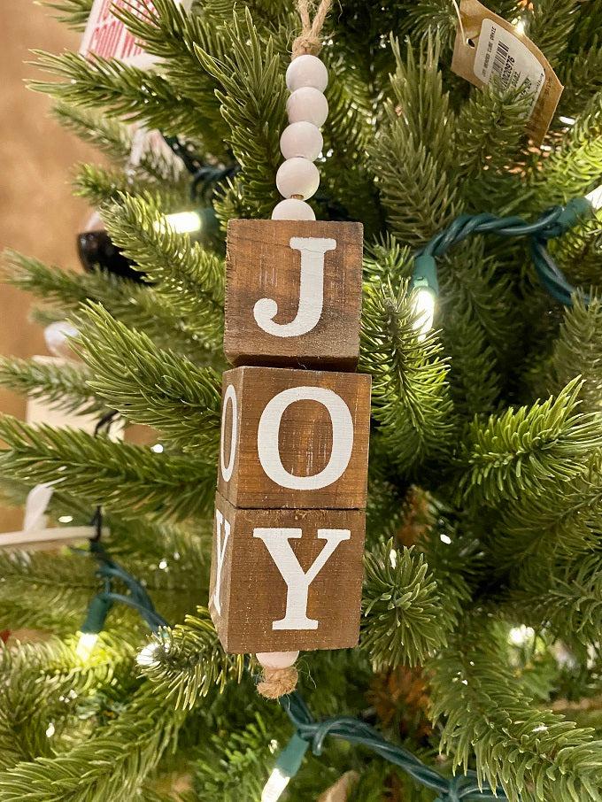 The Beaded Cube Ornament with the word JOY. The natural coloured cubes are printed with a single letter on all four sides and are moveable. Five white beads are at the top and one at the bottom of the ornament. 