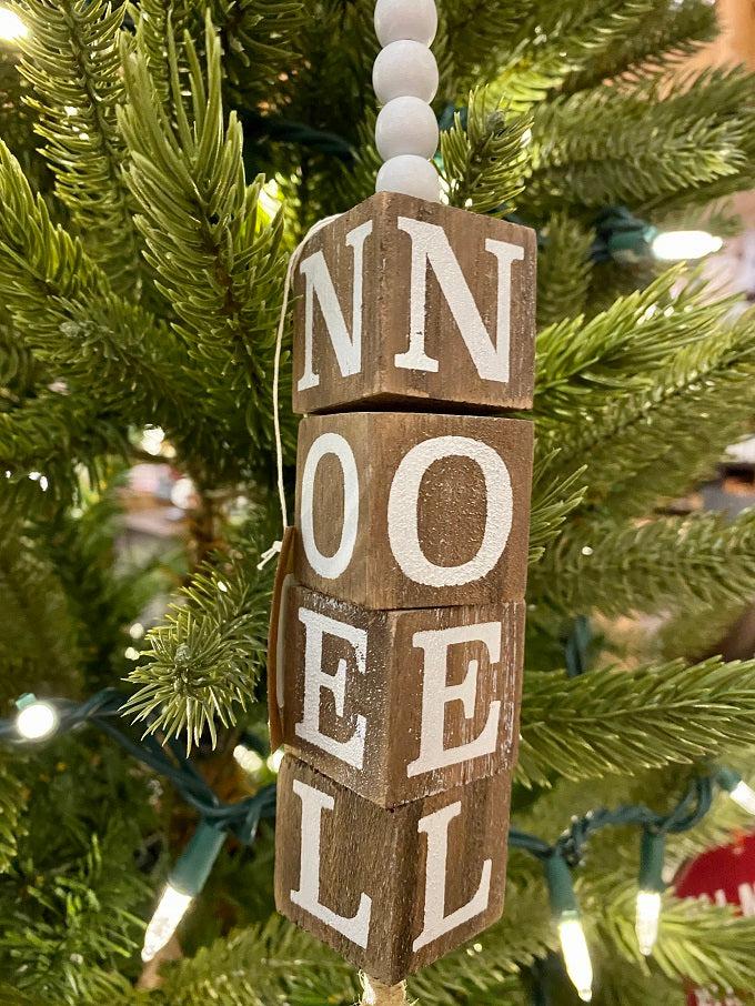 The Beaded Cube Ornament with the word Noel. The natural coloured cubes are printed with a single letter on all four sides and are moveable. Five white beads are at the top and one at the bottom of the ornament. 