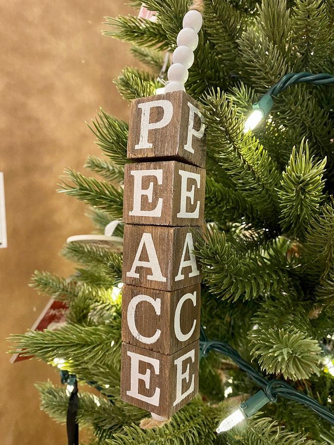 The Beaded Cube Ornament with the word Peace. The natural coloured cubes are printed with a single letter on all four sides and are moveable. Five white beads are at the top and one at the bottom of the ornament. 