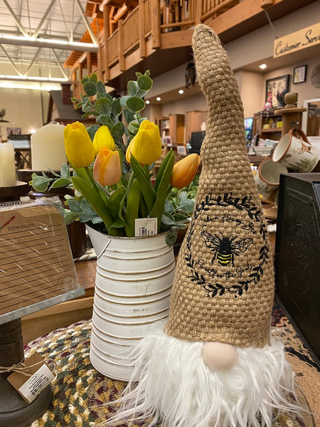The Honey Bee Gnome is a sitting gnome with a tall basket weave hat that is tan in colour.