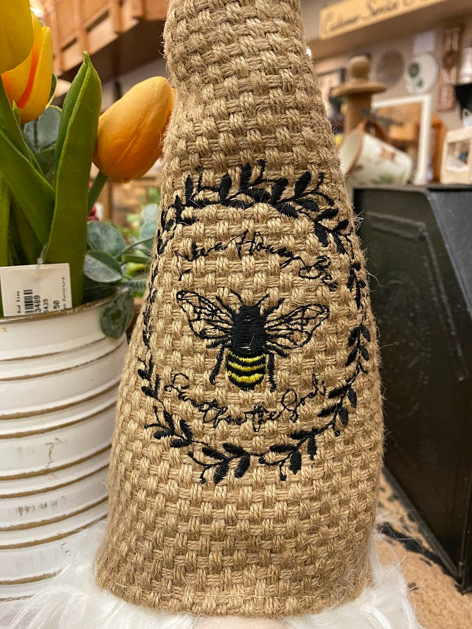 The Honey Bee Gnome is a sitting gnome with a tall basket weave hat that is tan in colour. A close up of the hat is shown with the embroidery featured. It is a honey bee and writing that says Like a honey bee, sweet for the soul.
