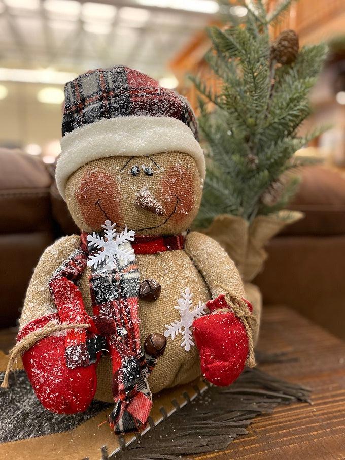 The Happy Burlap Snowman has a red and black plaid hat, is dusted with white snow and is big and round. His face has a big smile and rosy cheeks and there are a few big white snowflakes on his body. 