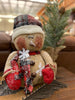 The Happy Burlap Snowman has a red and black plaid hat, is dusted with white snow and is big and round. His face has a big smile and rosy cheeks and there are a few big white snowflakes on his body. 