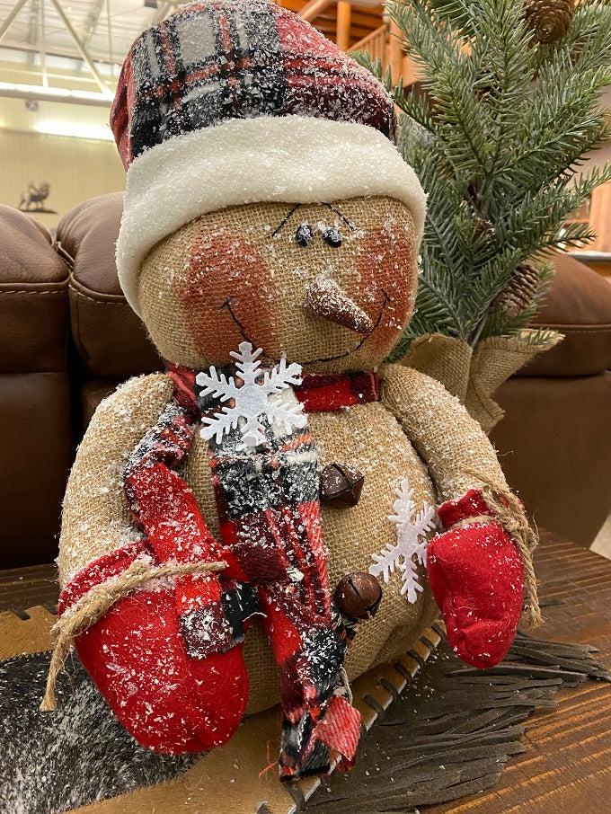 The Happy Burlap Snowman has a red and black plaid hat, is dusted with white snow and is big and round. His face has a big smile and rosy cheeks and there are a few big white snowflakes on his body. 