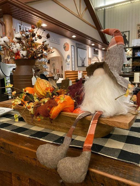 Pictured is a gnome with long dangle legs that will hang loosely. He has a plaid and brown hat with a big brimg and a full bushy beard that wraps around his whole body.  He is sitting in a wood bowl with fall gourds.