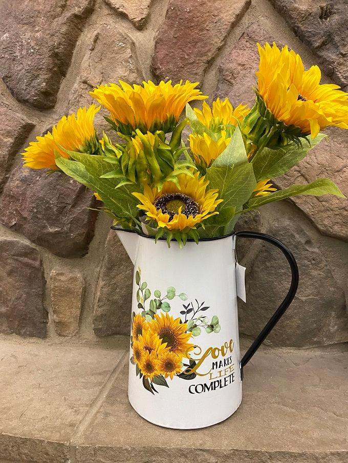 A white enamel pitcher is filled with beautiful sunflowers. The pitcher is white enamel, and has a black slim handle, and black painted trim around the top and spout. The picture on the side is of a bouquet of sunflowers and says Love makes life Complete.