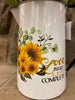 A close up of a white enamel pitcher. The picture on the side is of a bouquet of sunflowers and says Love Makes Life Complete..