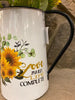 A close up of a white enamel pitcher. The picture on the side is of a bouquet of sunflowers and says love makes life complete..