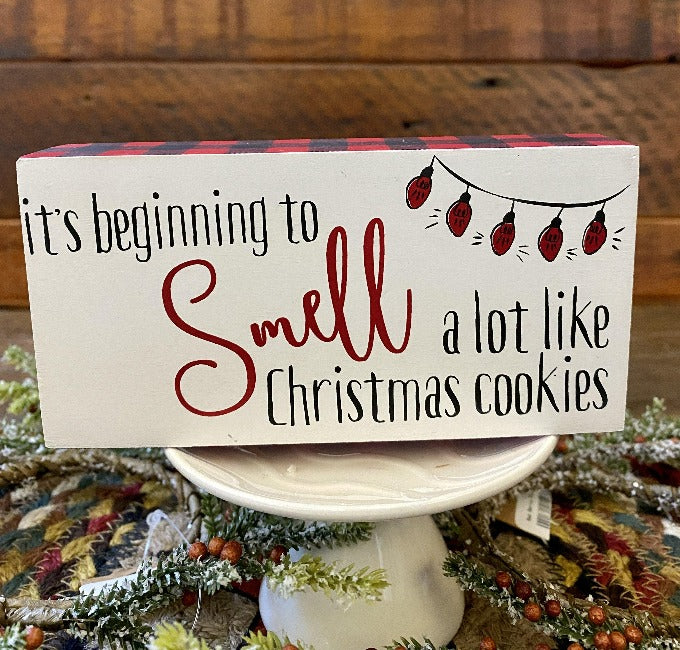 It's beginning to smell a lot like Christmas cookies block sign has a white background, red and black checked edge and It's beginning to smell a lot like Christmas cookies . It has a picture of christmas lights in the top corner. 