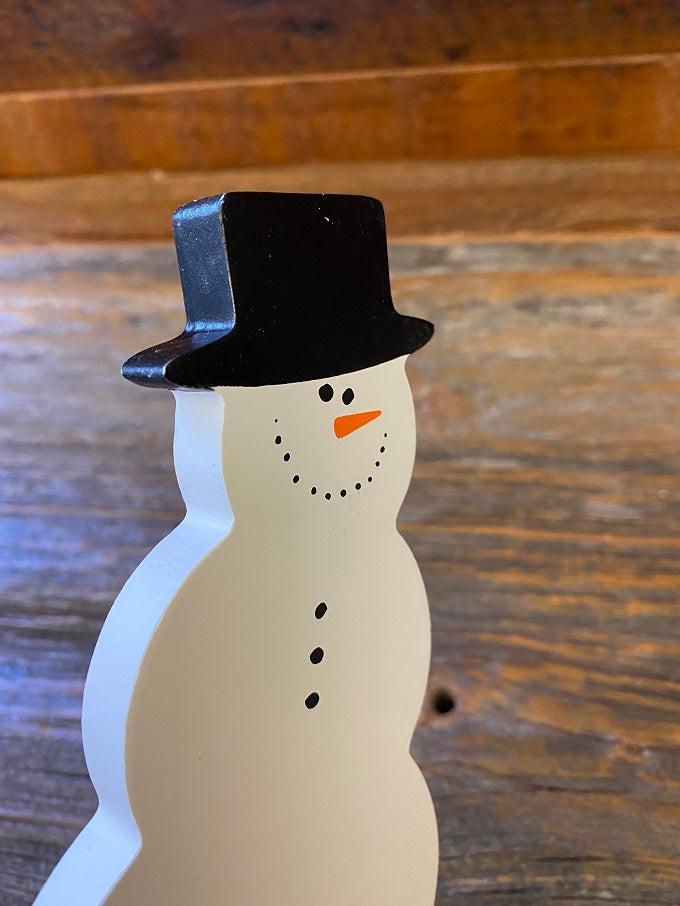 This is a wooden snowman cut out, so shaped like a snowman. It is painted white and its painted features include a black hat, carrot nose, coal for his eyes, nose and buttons.