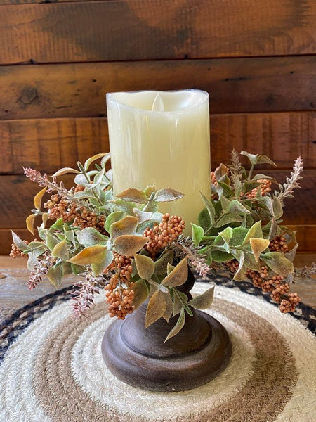 The pumpkin autumn harvest candle ring is shown here with a candle and candle holder. The ring has lots of greenery and as accents tiny orange beads clustered together with off white flowers.