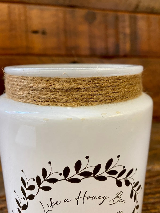 A white glass Honey Bee Jar with a image of a bee surrounded by a black vine of leaves and the words Like a Honey Bee Sweet for the Soul. The neck of the jar is wrapped in jute.