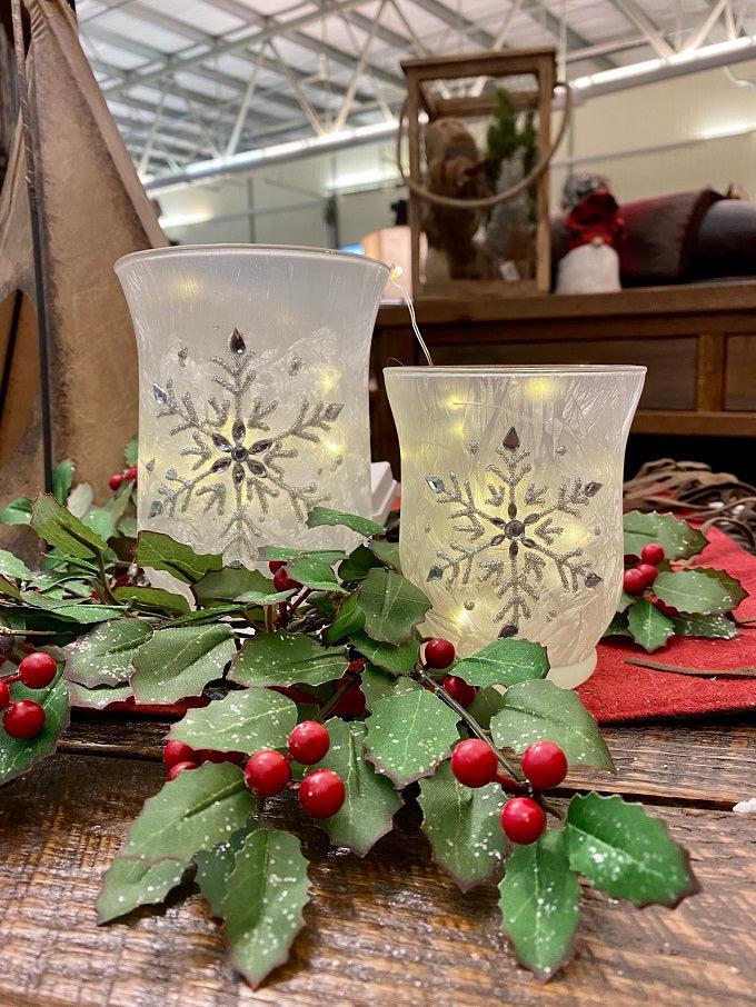 Frosted white glass hurricane shaped jars, small at bottom with a wider opening, have a snowflake pattern in silver on the outside. Two jars are shown. 