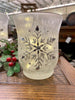 A single frosted white glass hurricane jar have a snowflake pattern in silver on the outside. 