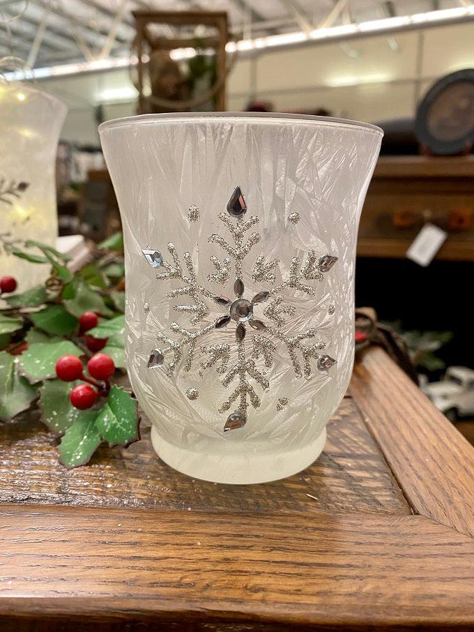 A single frosted white glass hurricane jar have a snowflake pattern in silver on the outside. 