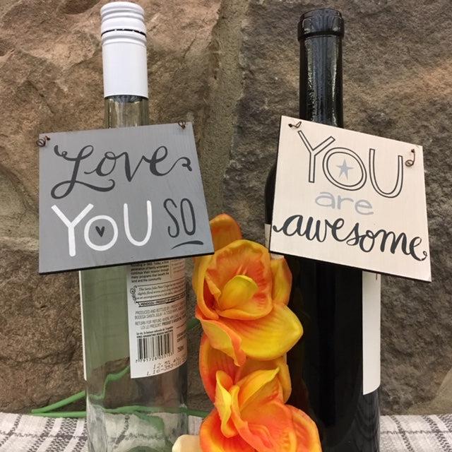 Two wine bottles with You wooden wine tags hanging from the neck of the bottle. One tag is dark gray and says love you soon it in white and black lettering. The other tag is a cream colour with the words You are awesome in gray and black lettering. The farmhouse wine tags make great gifts. You could write on them with a sharpie and give it as a gift with a bottle of wine. 