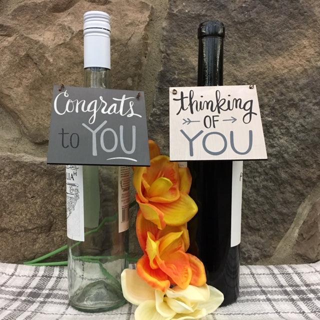 Two wine bottles with You wooden wine tags hanging from the neck of the bottle. One tag is dark gray and says Congrats to you it in white and black lettering. The other tag is a cream colour with the words Thinking of you in gray and black lettering. The farmhouse wine tags make great gifts. You could write on them with a sharpie and give it as a gift with a bottle of wine. 