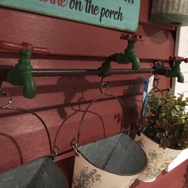 A top down look at the Three vintage distresssed metal half buckets with the word garden on the side hang from a pipe by their metal handle. Above each bucket is a green and red vintage faucet. The faucet spout is green and the tap is red. The metal buckets are distressed white in colour.
