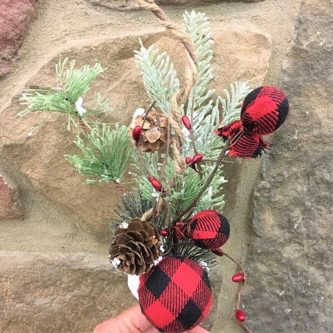 A pick made up of snowy Christmas greens, and round red and black check fabric coloured balls, pine cones and jute rope.