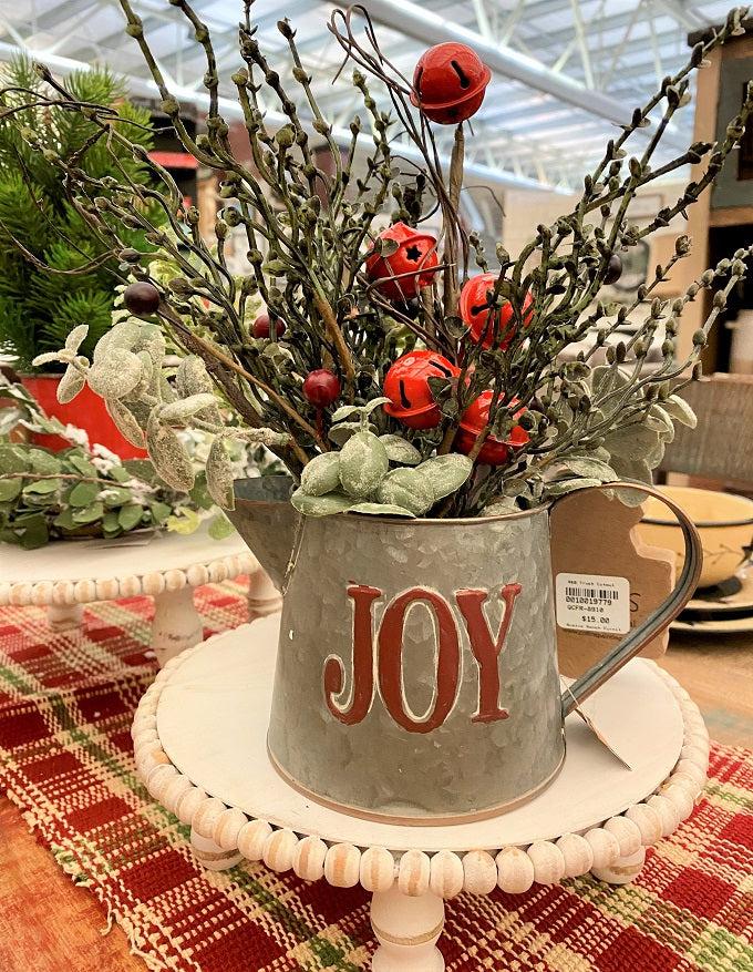 A galvanized filled with floral that says Joy on the outside. The bucket is full of green stems including the red bell pick.