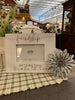A metal daisy is shown with a white picture frame. The daisy has spikey petals and there are four layers of them. It is made of galvanized metal. The centre of the flower has raised metal pieces coming together for a pretty centre. There is white distressed paint on the edges of the petals.