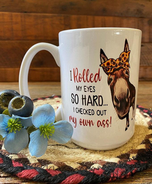 A white ceramic mug with a brown donkey, cartoon style, with a yellow and brown polkadot bow. Beside the donkey are words in red and black that say I rolled my eyes so hard I checked out my own ass.