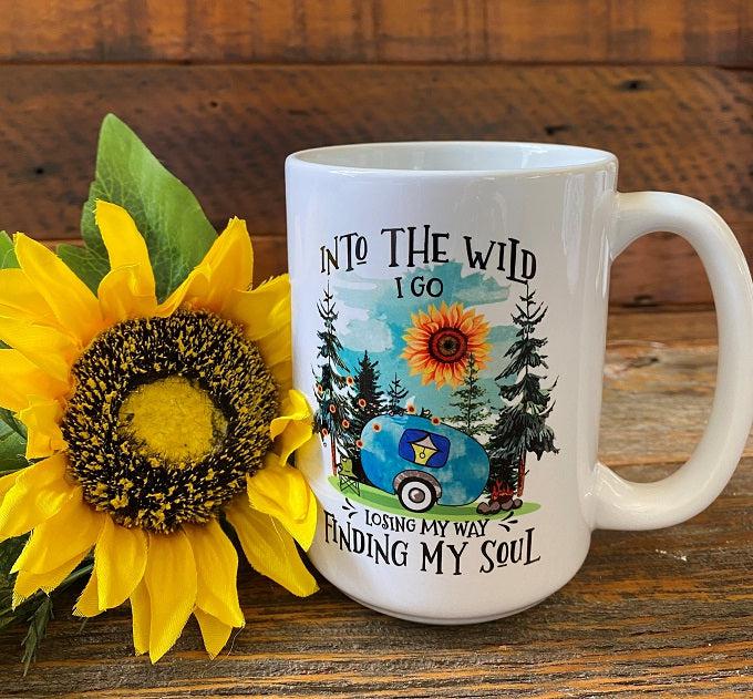 A White ceramic coffee mug that is imprinted on both sides with a sentiment that says Into the wild I go, losing my way finding my soul. These words are around a camping scene picture of a blue camper, with a sunflower sun and pine trees.
