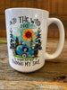 A White ceramic coffee mug that is imprinted on both sides with a sentiment that says Into the wild I go, losing my way finding my soul. These words are around a camping scene picture of a blue camper, with a sunflower sun and pine trees.