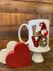 The Love Gnome mug is shown with other valentine decor. The gnome on the mug has a red and black buffalo plaid hat. The V of Love is also Red and Black Buffalo plaid and the L and E are leopard print