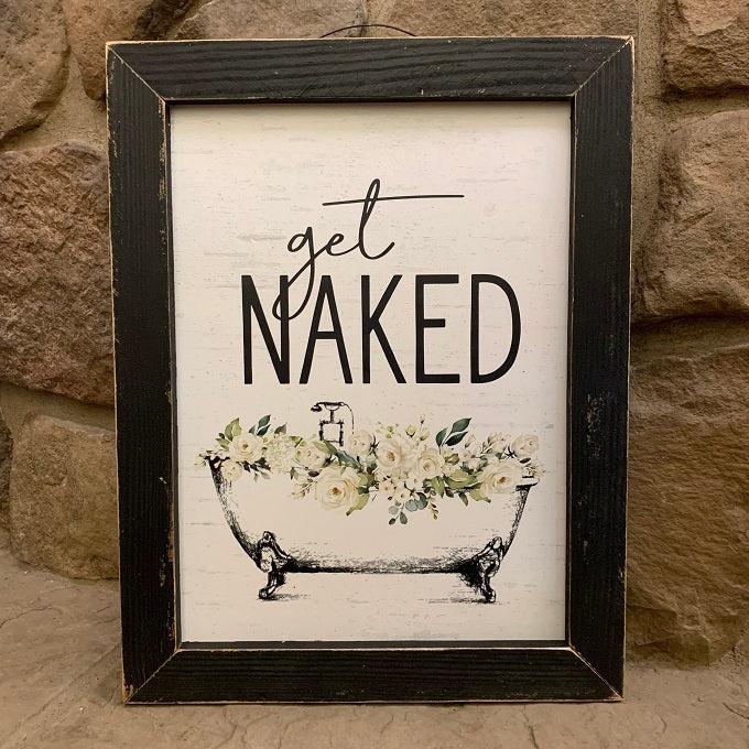 This bathroom print has a vintage bath tub filled with white flowers and greens and it reads, "Get Naked" above the tub.  The bathtub is white and filled with yellow-toned flowers. it is a claw foot bathtub. The words get naked are in black. It has a black wooden distressed frame and comes with a vintage wire hanger for ease of hanging.
