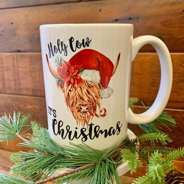 A white ceramic mug printed on both sides that has a picture of a Highland cow with a red santa hat.  It is surrounded by the words Holy Cow It's Christmas.