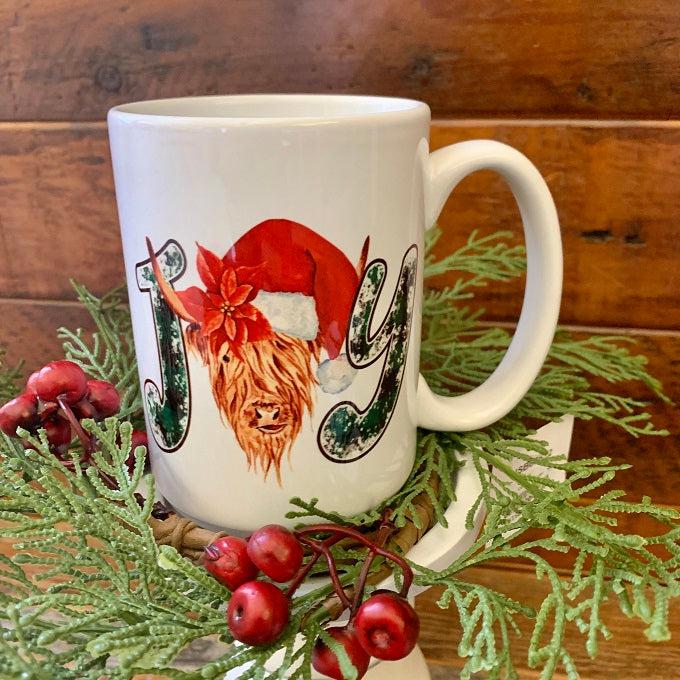 A highland cow with a a red santa hat is the 'O" in the word JOY on this white ceramic mug.